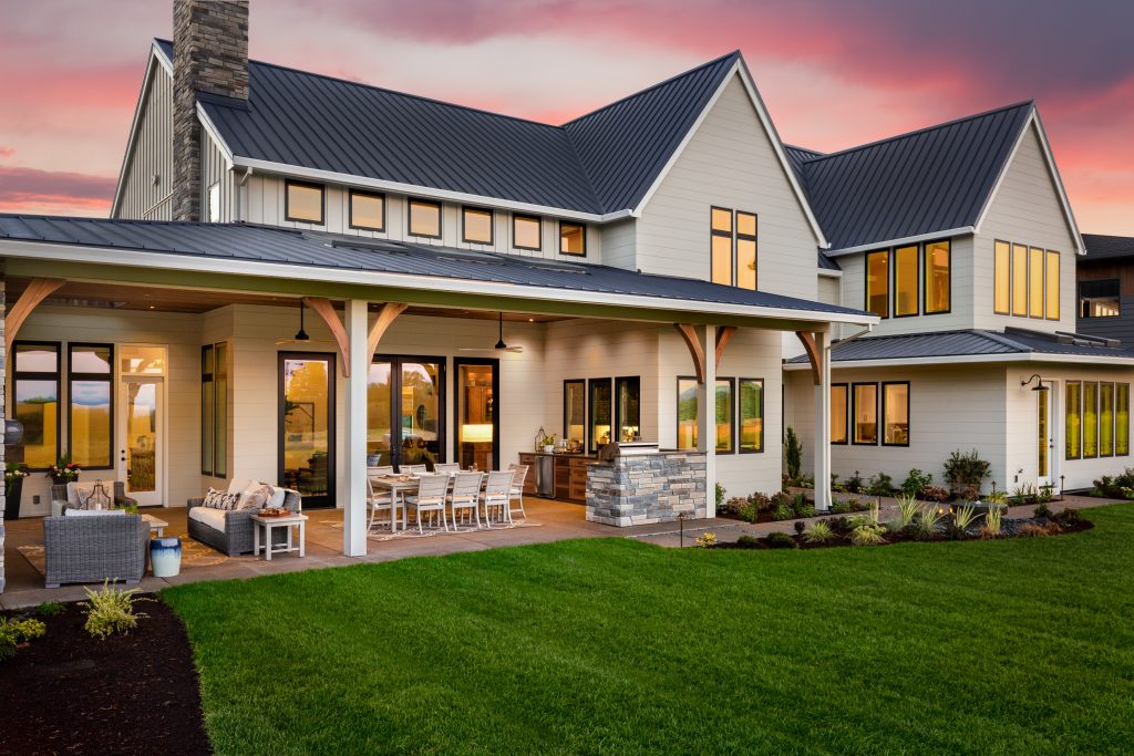 New luxury home exterior at sunset. Features large covered patio with barbecue, outdoor kitchen, and furniture